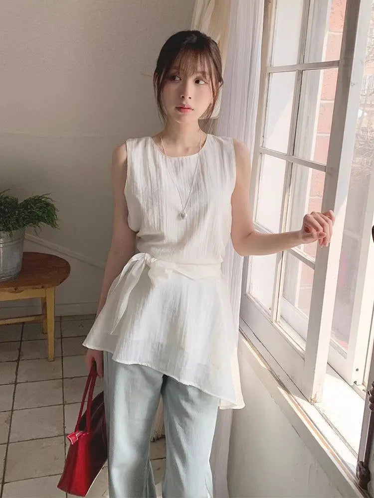 A woman stands by a window wearing a sleeveless white top with a tie waist and light blue pants, holding a red handbag in a bright indoor setting.