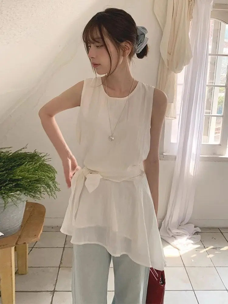 A woman wearing a light gray sleeveless linen wrap top with a tie-neck detail, paired with white pants, holding a metallic silver handbag in an indoor setting.