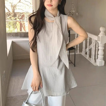 A woman wearing a light gray sleeveless linen wrap top with a tie-neck detail, paired with white pants, holding a metallic silver handbag in an indoor setting.